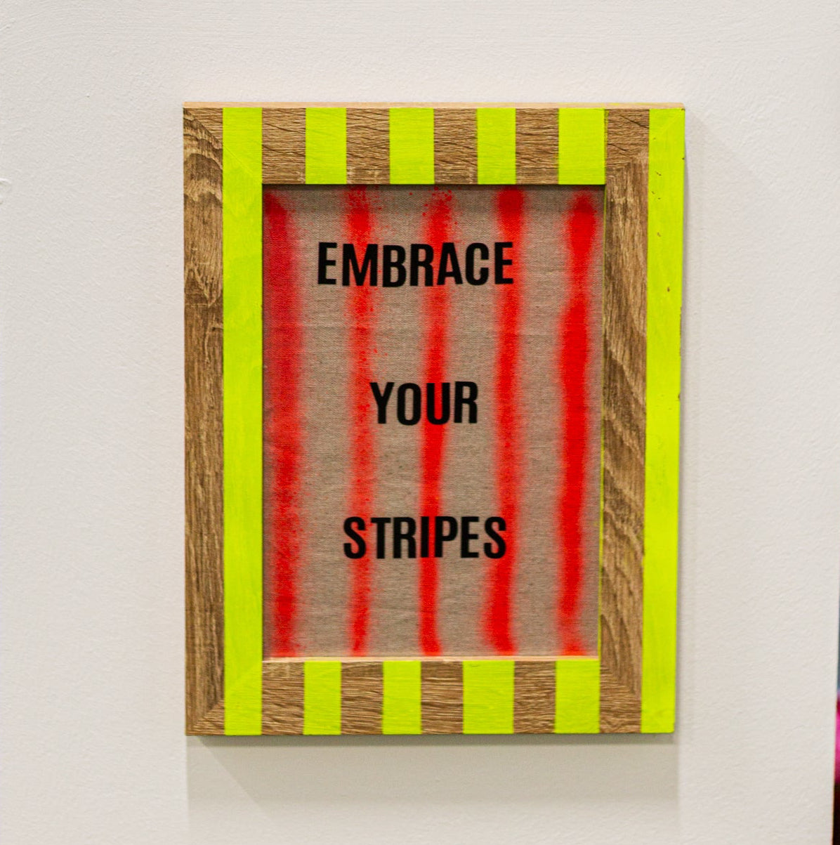Hand-painted stripe photo frame in neon with  motivational sign reading 'Embrace Your Stripes' on a white wall 
