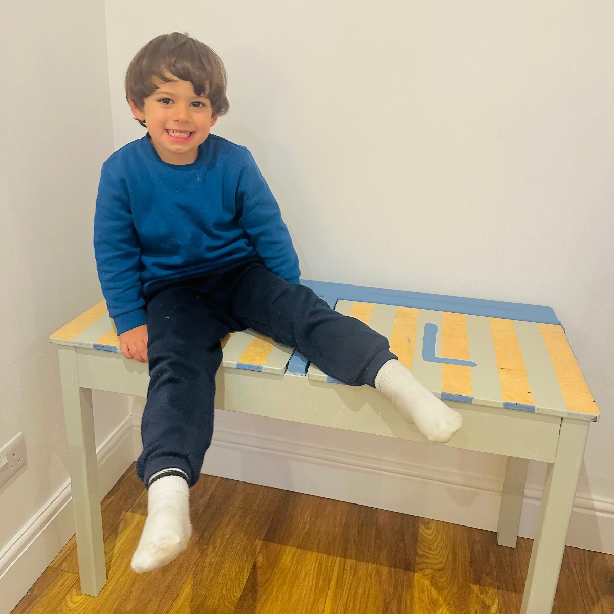 Child sitting on a small table with a colorful top in a room with wooden flooring and white walls.