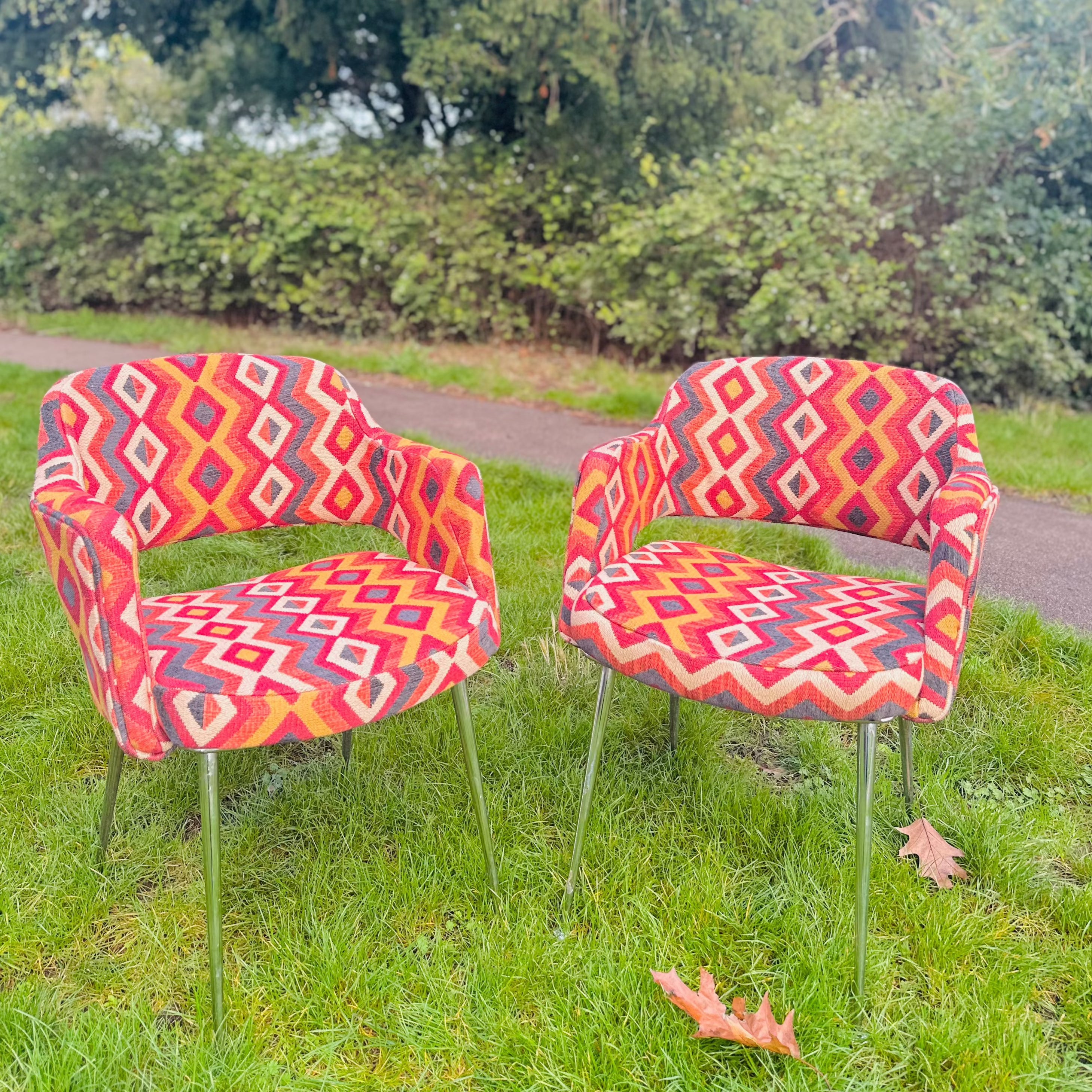 Two upholstered retro dining chairs with a geometric pattern, placed on a grassy area with trees in the background.