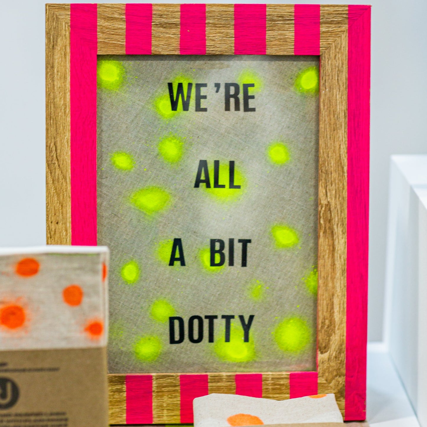 handprinted striped eco wooden frame with 'WE'RE ALL A BIT DOTTY' text and colorful accents on a white background.